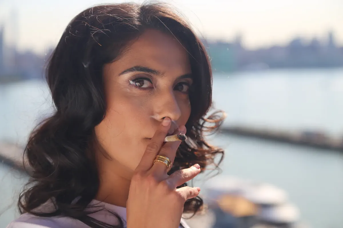 Woman with dark hair smoking near a waterfront with city skyline in Hoboken, NJ.