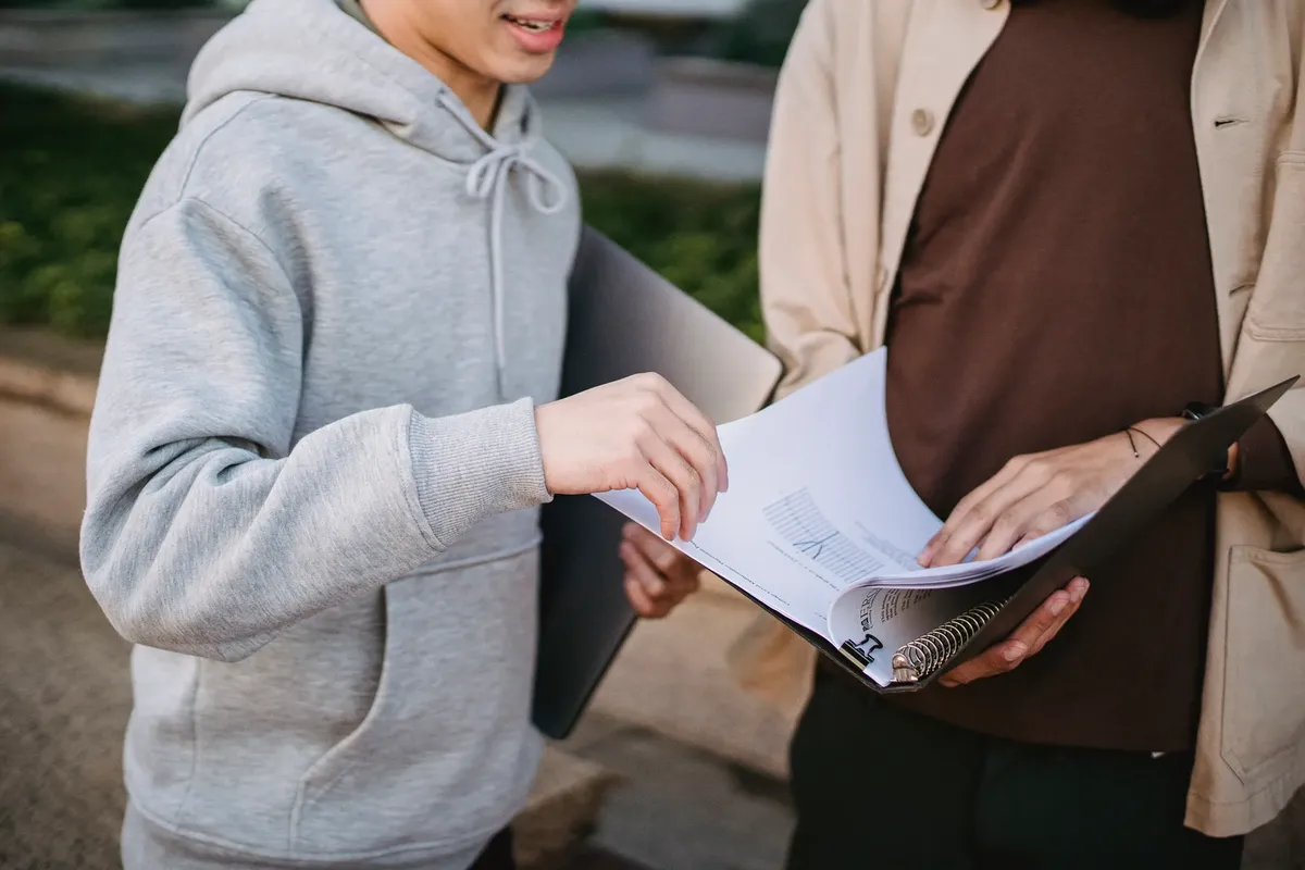 Crop male students in casual wear reading learning material in folder while standing together in university park