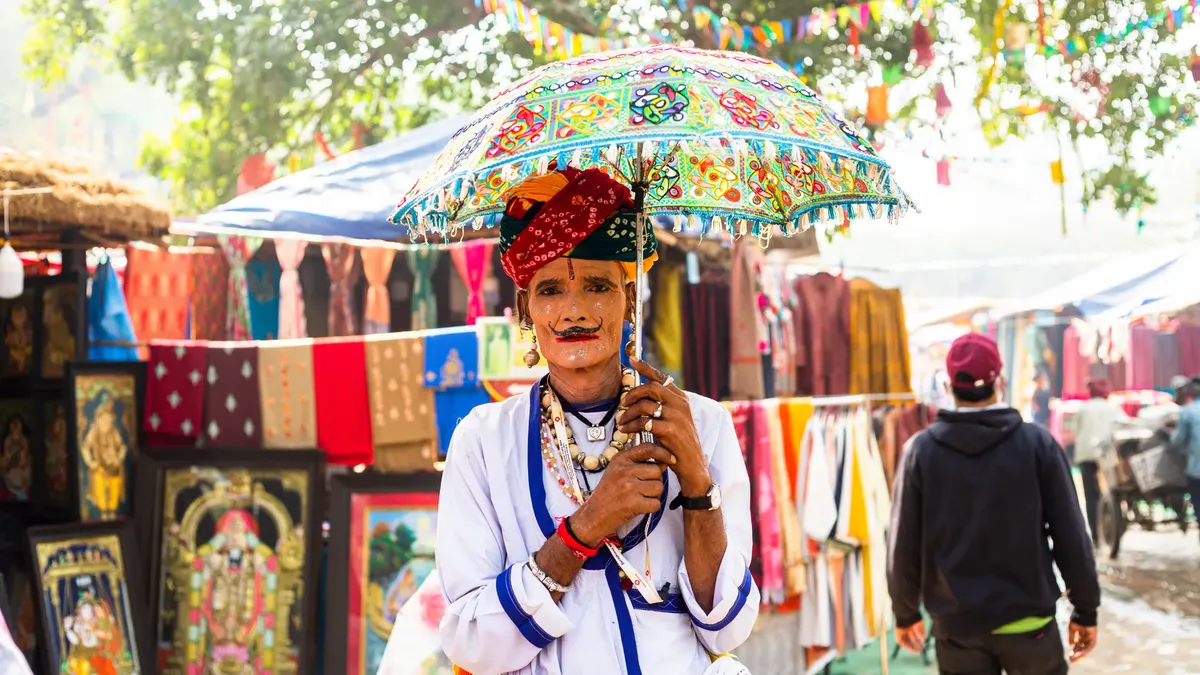 Man in traditional Indian clothing with face paint holding a colorful umbrella at a market.